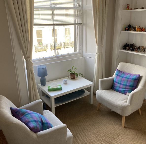 A bright room with 2 comfy chairs set opposite each other by a large window, there's a table nearby with tissues on it, behind one of the chairs is a bookcase full of figurines